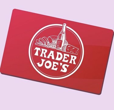 Κόκκινη δωροκάρτα του Trader Joe σε ροζ φόντο. Το λογότυπο του Trader Joe βρίσκεται στη μέση της κάρτας με λευκό κείμενο και μια εικόνα με φέτες ψωμιού, σταφύλια, ένα μπουκάλι κρασί και τυρί, επίσης σε λευκό.