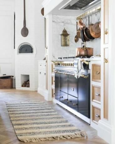 бяла кухня в френски стил в кънтри с черна печка и килим, медни тенджери, висящи отгоре