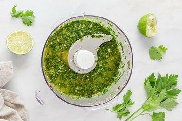 Una ciotola per robot da cucina riempita con la salsa chimichurri. È circondato da lime e coriandolo.