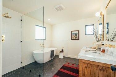 Didelis vonios kambarys su berėmiu dušu, medinėmis ir marmurinėmis kriauklėmis, pilkomis keraminėmis grindų plytelėmis ir balta vonia