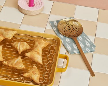 Samosa sopra una griglia di raffreddamento dorata in una teglia da forno color curcuma accanto a un cucchiaio forato su un tovagliolo blu e bianco.