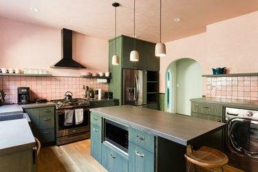 Keittiö, jossa on vaaleanpunaiset seinät, vihreät kaapit ja hyllyt, harmaat tiskit, vaaleanpunainen takalevy ja kelloripustimet.