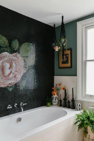 hvitt badekar med svart vegg med rosemosaikk, hengende planter fra svart makrame, hvite fliser på veggene, planter rundt badekaret