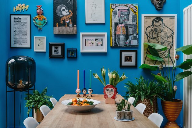 paredes azuis brilhantes em uma sala de jantar com arte da parede da galeria e cadeiras eames moldadas em branco em uma mesa de jantar de madeira