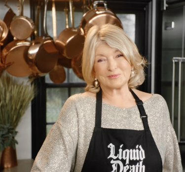 Een glimlachende Martha Stewart in een keuken met een zwart Liquid Death-schort over een beige trui.