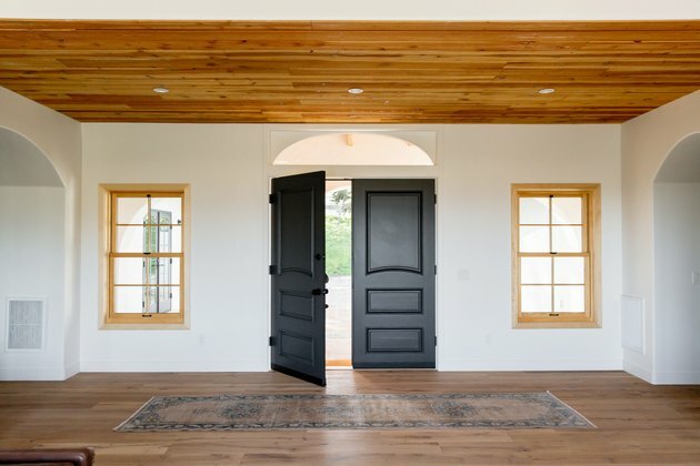 projeto de casa ecologicamente correto com hall de entrada de uma casa de estilo espanhol com portas duplas de madeira escura, uma das quais aberta, em uma parede branca. Duas janelas com molduras de madeira clara de cada lado da porta. O piso e o teto são ambos de madeira. Há um tapete na frente da porta.