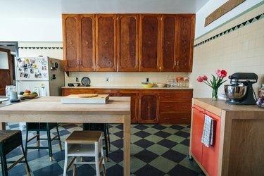 Küche mit Holzschränken und Esstisch, Hockern, Wänden mit beigen Fliesen und dreieckigen Akzentfliesen, einem tragbaren Schrank mit rosa Türen.