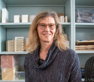 La arquitecta Helen Bronston, con cabello rubio hasta los hombros, usa un par de anteojos de color marrón claro y una camisa azul oscuro con cuello vuelto frente a una biblioteca azul claro.