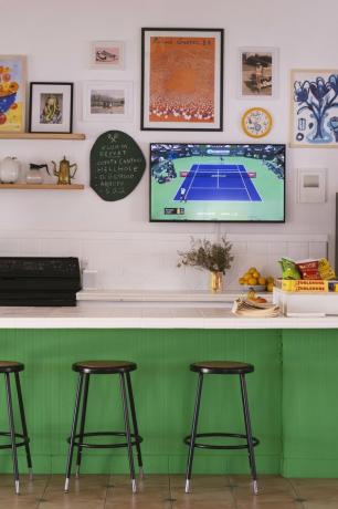 Cozinha contemporânea com estrutura de bar com laterais de azulejos verdes, bancada branca, bancos pretos, TV de tela plana e parafernália esportiva pendurada na parede rosa