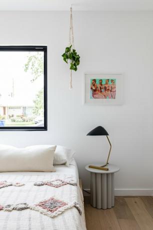 Quarto com mesa lateral com abajur, cama branca com travesseiro e cobertor acolchoado, piso de madeira, parede branca com janela, planta suspensa e pintura