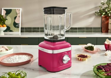 Hibiscus KitchenAid -sekoitin lepää keittiön tiskillä matalan jauholautasen ja raakajauhelautasen keskellä. kanaa, kaksi vihreää ja punaista chiliruokaa, kultainen punaisia ​​mausteita ja ruokalaji sipulia ja vihreää paprikat.