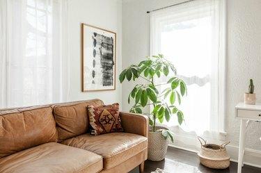 Svijetla dnevna soba s podom od tamnog drveta. Smeđa kožna sofa s tradicionalnim jastukom, apstraktnom umjetnošću, biljkom drveća i košarom.