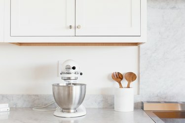 Armadietti bianchi sopra un bancone in marmo. Sul bancone è presente un robot da cucina KitchenAid bianco con ciotola in acciaio inox. Accanto al mixer c'è un portautensili bianco con una pinza per insalata in legno.