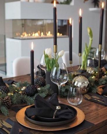 Mesa con platos negros, candelabros y piñas oscuras y manteles dorados y portavelas