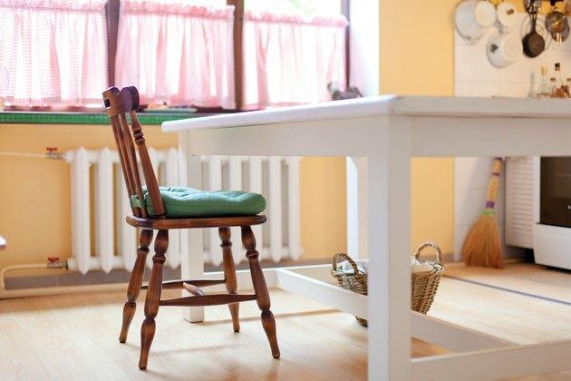Kuhinjski prizor z belo mizo, lesenim stolom in zeleno blazino. Prijeten dom s košarico wisker na tleh in baterijo za ogrevanje pod velikim oknom.