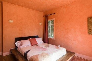 غرفة نوم بجدران برتقالية وشمعدانات جدارية بيضاء ووسائد ووسائد برتقالية وستائر برتقالية وأرضية خشبية داكنة.