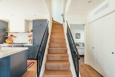 Laiptai su medinėmis grindimis ir juodais bėgiais šalia virtuvės su mėlynomis spintelėmis ir marmuriniais stalviršiais