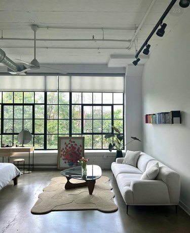 Una sala de estar con grandes ventanales, un sofá blanco y una alfombra beige debajo de una mesa de café.
