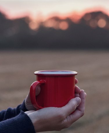 Henkilö, jolla on punainen emalimuki ulkona katsellessaan auringonlaskua pellolla.