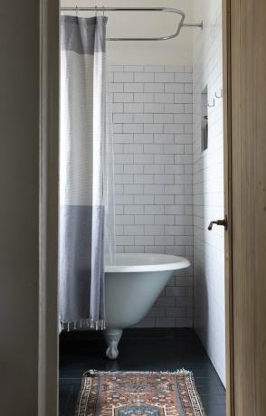 um banheiro com piso de metrô, um pequeno tapete e uma banheira com pés com uma cortina cinza e branca