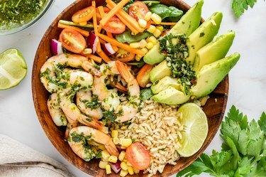 Una ciotola di legno piena di gamberetti chimichurri. riso, fette di avocado, lime e un mix di pomodorini, mais e carote. Sul piano di lavoro in marmo circostante, c'è uno spicchio di lime, un fascio di coriandolo e una ciotola di salsa chimichurri.