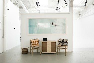 सफेद दीवारों वाली आर्ट गैलरी जिसमें बड़े प्रिंट, तकिये वाली कुर्सियाँ और कंक्रीट के फर्श पर लकड़ी की कैबिनेट है