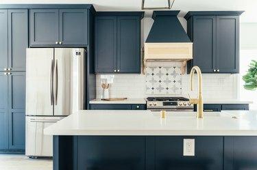 Mørkeblå kjøkkenskap med gullknotter. Et sølvfarget fransk kjøleskap. Utsmykkede fliser ved komfyrtopp. Kjøkkenøy med gull servantkran.