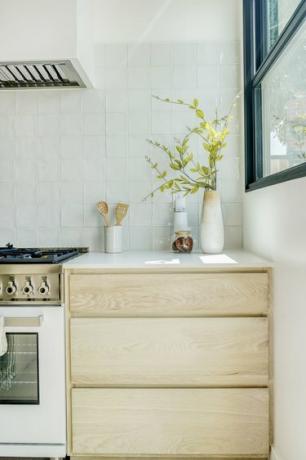 Un mueble de cocina de madera clara con jarrones de recortes de flores y un jarrón con utensilios de madera, todo junto a una estufa