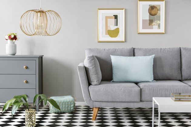 Lys blå pude placeret på grå sofa i lyst stueindretning med linoleumgulv i betræk, friske blomster i vase på skab to plakater hængende på væggen og guldlampe
