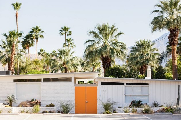 exterior da casa branca em meados do século Palm Springs casa com porta laranja rodeada por palmeiras