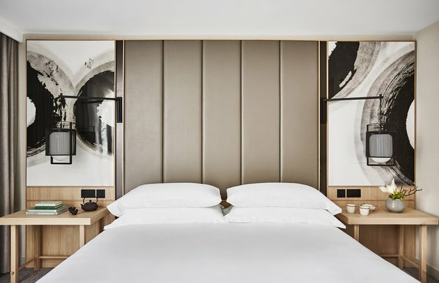 Cama king-size com roupa de cama branca e travesseiros com cabeceira com painéis de tecido na altura do teto, mesas laterais de madeira e pinturas acima