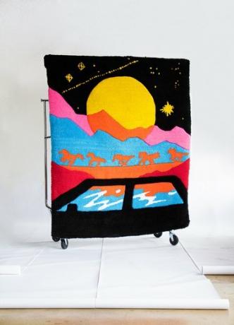 Stačiakampis kilimas, vaizduojantis kelionės sceną su juodu automobiliu ir įvairiaspalvį kalnų peizažą su oranžiniais arkliais ir dideliu mėnuliu su žvaigždėmis juodame danguje.