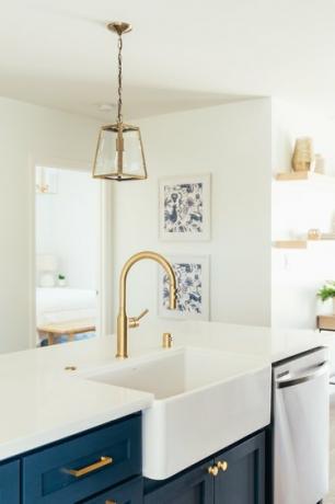Guld vandhanevask i en køkkenø med mørkeblå skabe og hvid bordplade. Guld lanterne pendel.