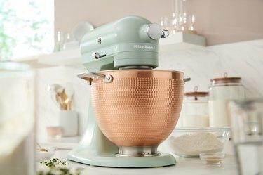 Pehmeän vihreä KitchenAid Blossom -telinesekoitin pronssisella kulholla valkoisella työtasolla.