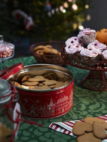 Punainen tölkki täynnä keksejä vihreällä painetulla pöydällä, takana muita makeisia, kuten vaaleanpunaisia ​​​​himmenkuppikakkuja ja keksikulhoja.