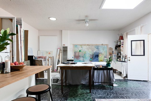 Idéias de garagem em Galeria de artistas com paredes brancas, com grande pintura fixada em uma parede, próximo a estantes com material de pintura e pequena ilha de cozinha com banquinhos marrons