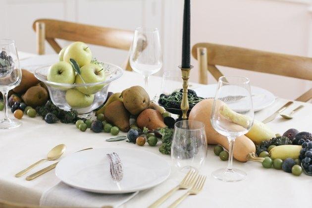 herfstfeestidee met tafelloper van producten, waaronder druiven, peren, kersen en pompoen loopt over de lengte van een tafel met een wit kleed en gouden bestek