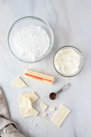 Ciotole di vetro piene di zucchero a velo e crema di formaggio, accanto a un panetto di burro, un cucchiaino di estratto di vaniglia e pezzi di cioccolato bianco su un piano di lavoro in marmo bianco.