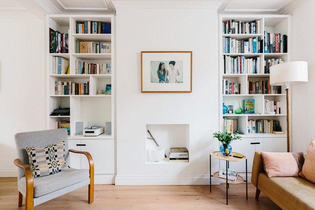 Como organizar uma estante de livros na sala de estar moderna com estantes, sofá de couro, cadeira, mesinha lateral e uma pequena lareira branca embaixo da imagem emoldurada pendurada