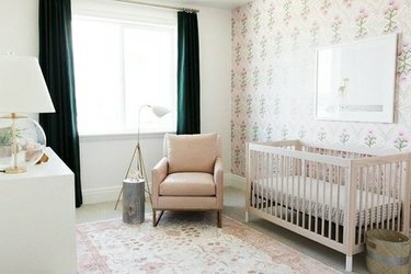 Детска стая с флорални тапети, тъмнозелени завеси, розов стол, флорален килим.
