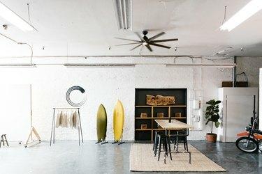 Индустриално студио с дъски за сърф, гардероб, дървена маса, растение, вентилатор на тавана и флуоресцентни лампи.
