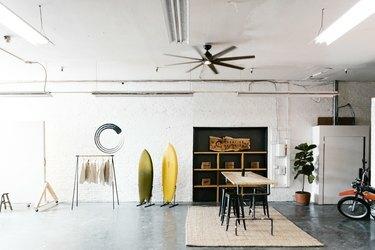 Un studio industriel avec des planches de surf, un placard, une table en bois, une plante, un ventilateur de plafond et des lampes fluorescentes.