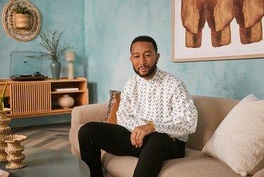 Musikeren John Legend sitter på en sofa iført svarte bukser og en hvit og svart skjorte med knapper.