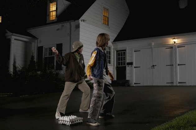 Due ragazzi (12-13) in piedi nel vialetto, lanciando uova a casa, notte, vista posteriore