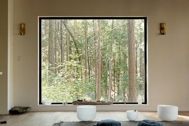 Большое панорамное окно с видом на лес и кристаллами. Нейтральный коврик с синими подушками и белыми поющими чашами для медитации.
