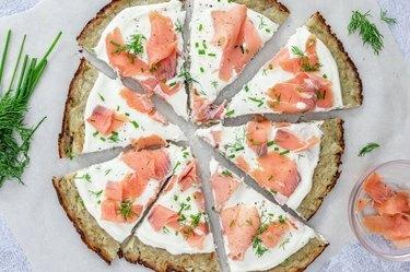 Ilmakuva Smoked Salmon Latke Pizzasta, jota ympäröi tuore tilli, ruohosipuli ja savulohen täytetty pieni lasikulho.