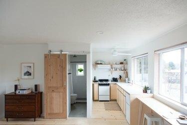 Vedere completă la studio cu pereți albi, dulap, ușă de baie din hambar, bucătărie cu rafturi deschise
