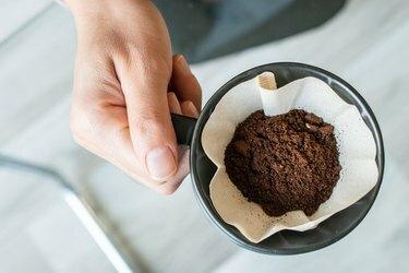 Apkarpytas vaizdas, kuriame barista laiko puodelį su malta kava viduje su popieriniu filtru, prieš jį lašinant.