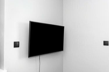 El televisor está colgado en la pared gris del dormitorio, los botones de las persianas son visibles.