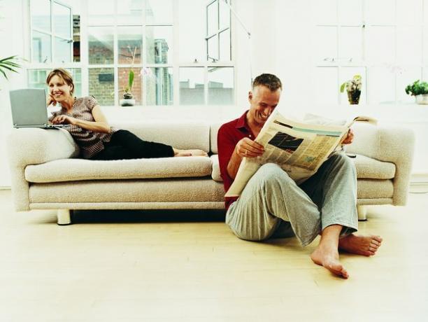 Verslo pora, sėdinti bute, esančiame gyvenamajame kambaryje, skaitant laikraštį ir naudojantis nešiojamu kompiuteriu